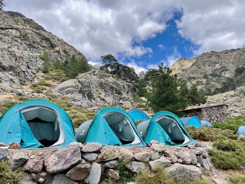 Une rangée de tentes de camping turquoise installées sur une terrasse en pierre dans un paysage de montagne rocheux sous un ciel partiellement nuageux.