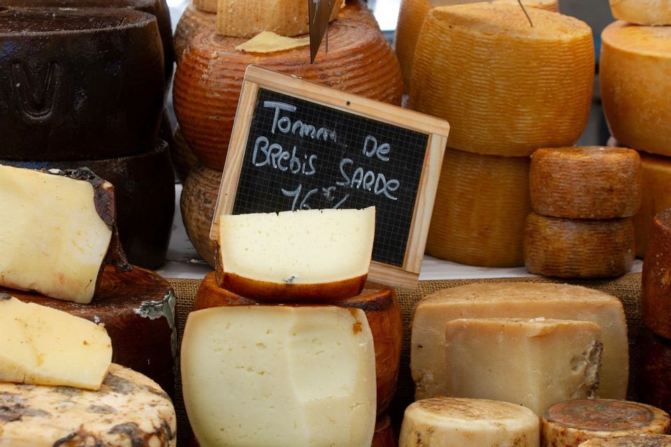 Différentes meules et portions de fromage exposées sur un étal de marché avec une ardoise indiquant 'Tomme de Brebis Sarde'.