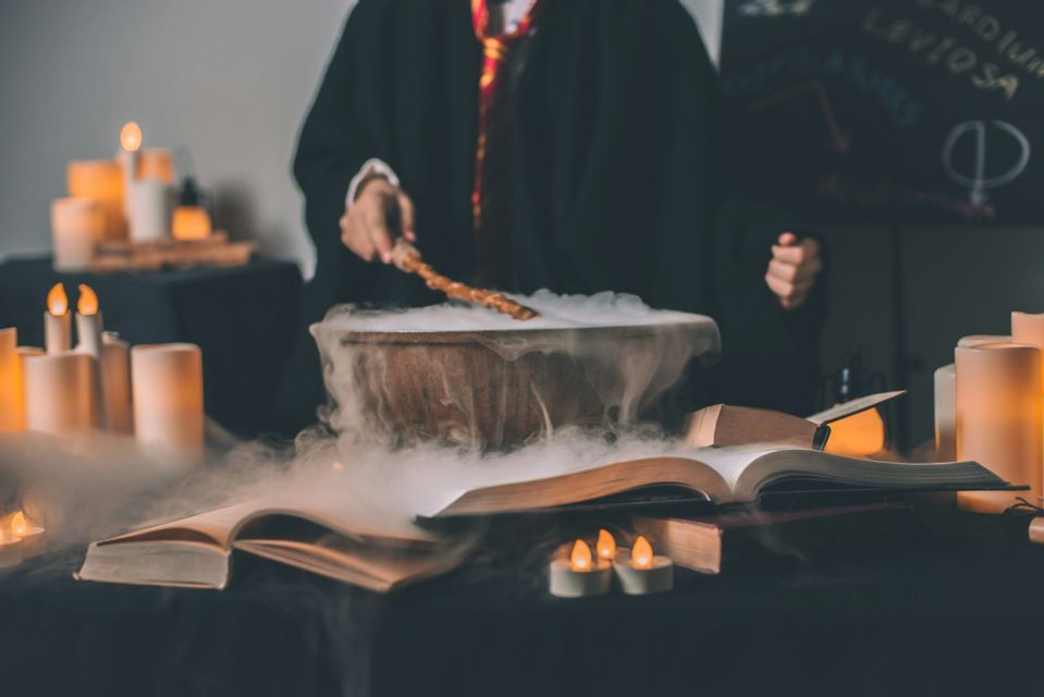 Una persona in una veste scura mescola un calderone fumante con una bacchetta magica, circondata da libri aperti e candele.