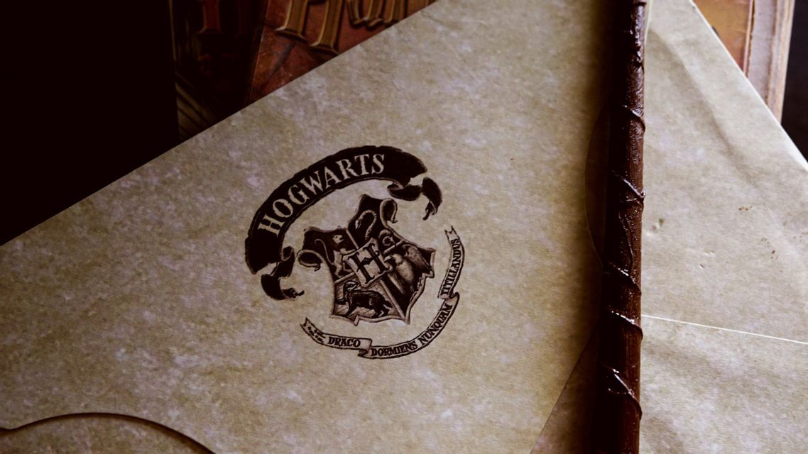 Un primo piano di una pergamena con lo stemma di Hogwarts stampato sopra, adagiata accanto a una bacchetta magica oscura.