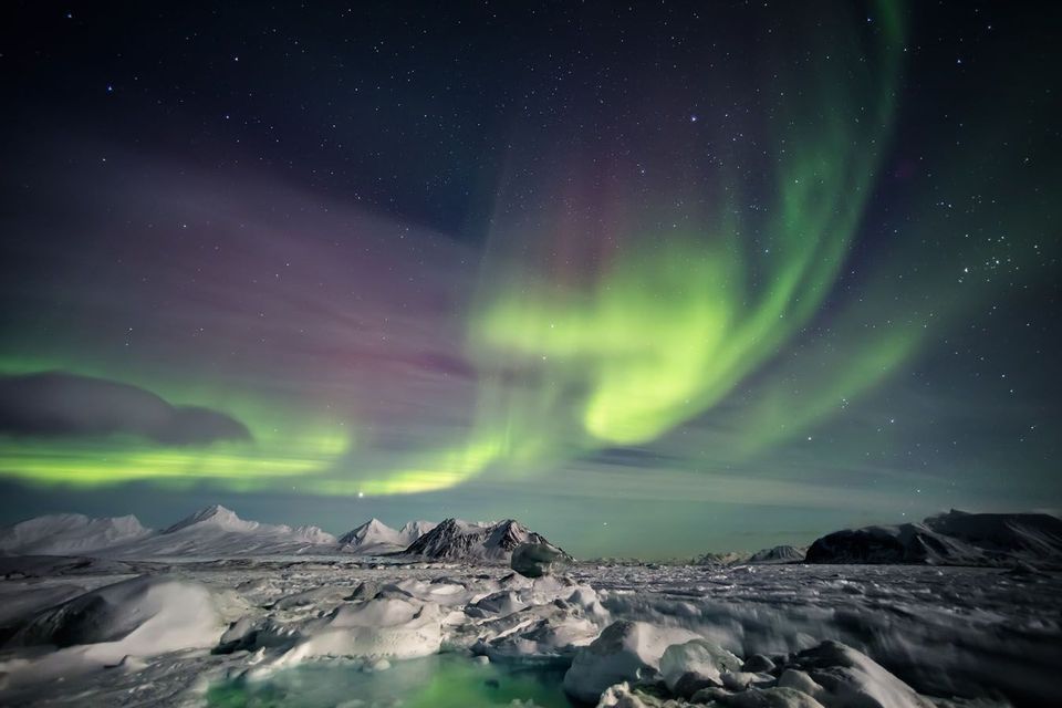 L'aurora boreale risplende verde e viola in un cielo notturno stellato sopra un paesaggio montuoso innevato con ghiaccio spezzato.