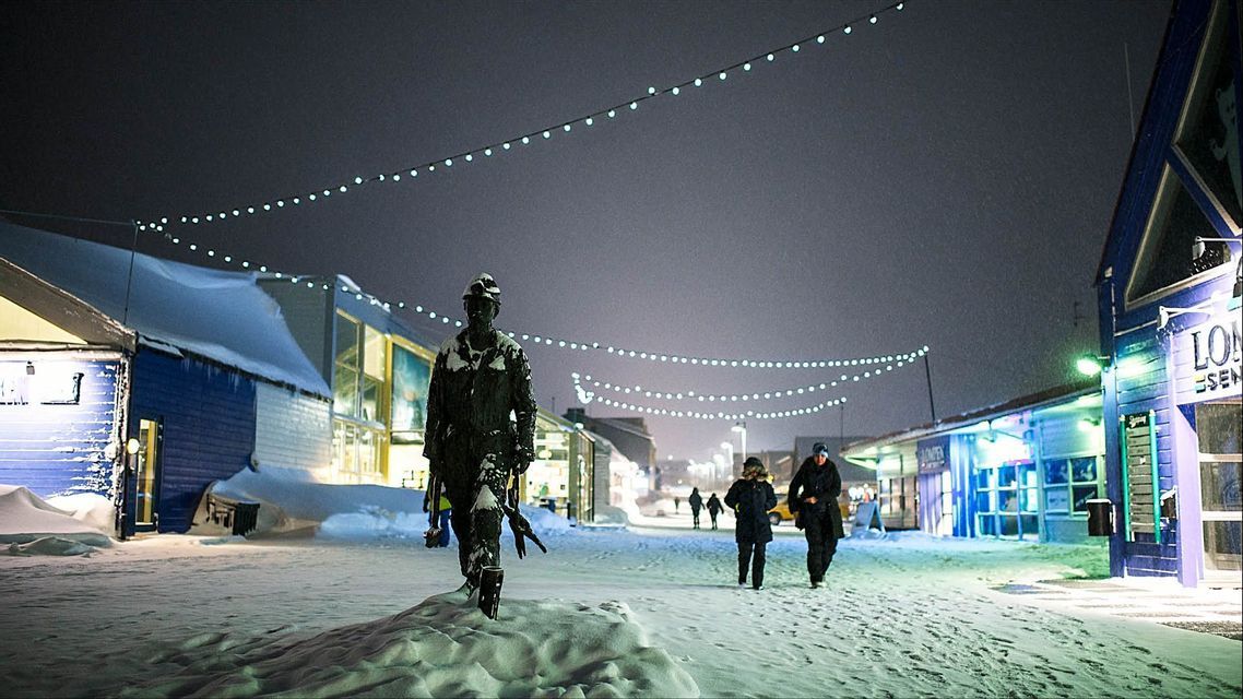 Di notte, una statua innevata di un minatore si staglia su una via cittadina, mentre le persone passeggiano sotto filari di luci.