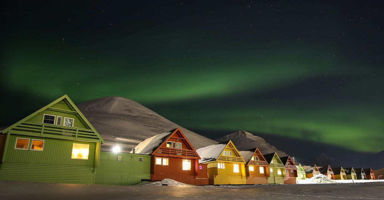 Una fila di case di legno colorate con finestre illuminate si trova nella neve sotto il bagliore verde dell'aurora boreale di notte.
