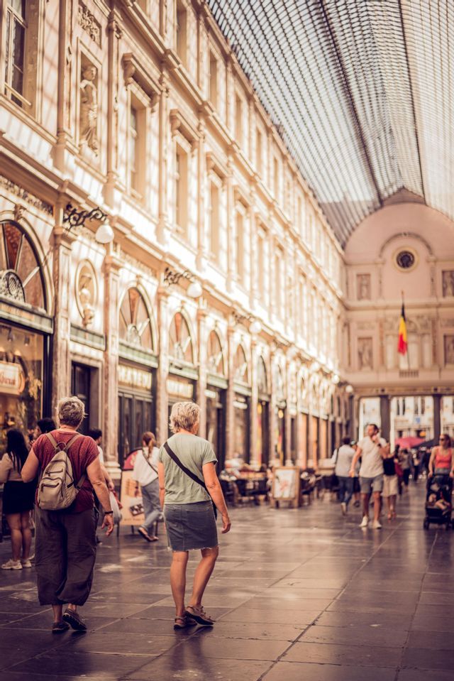 Deux femmes marchent de dos à travers une galerie marchande ornée et couverte de verre, remplie d'autres piétons.