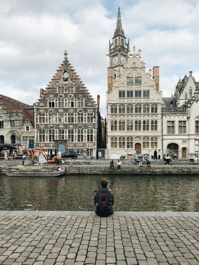 Une personne avec un sac à dos est assise sur un quai pavé, regardant de l'autre côté d'un canal des bâtiments historiques à pignons sous un ciel nuageux.