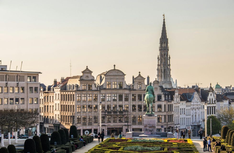 Une vue depuis un jardin bien entretenu d'une place de ville avec une statue équestre, des bâtiments historiques et une haute flèche contre un ciel brumeux.