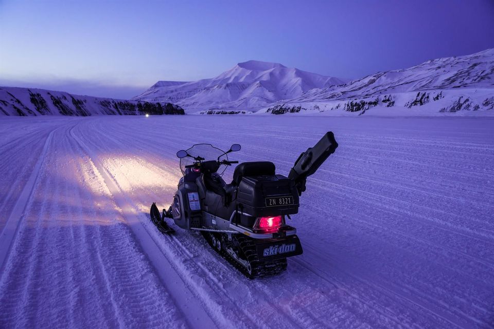 Una motoslitta con i fari accesi è parcheggiata su una pista innevata al crepuscolo, con vaste montagne coperte di neve sullo sfondo.
