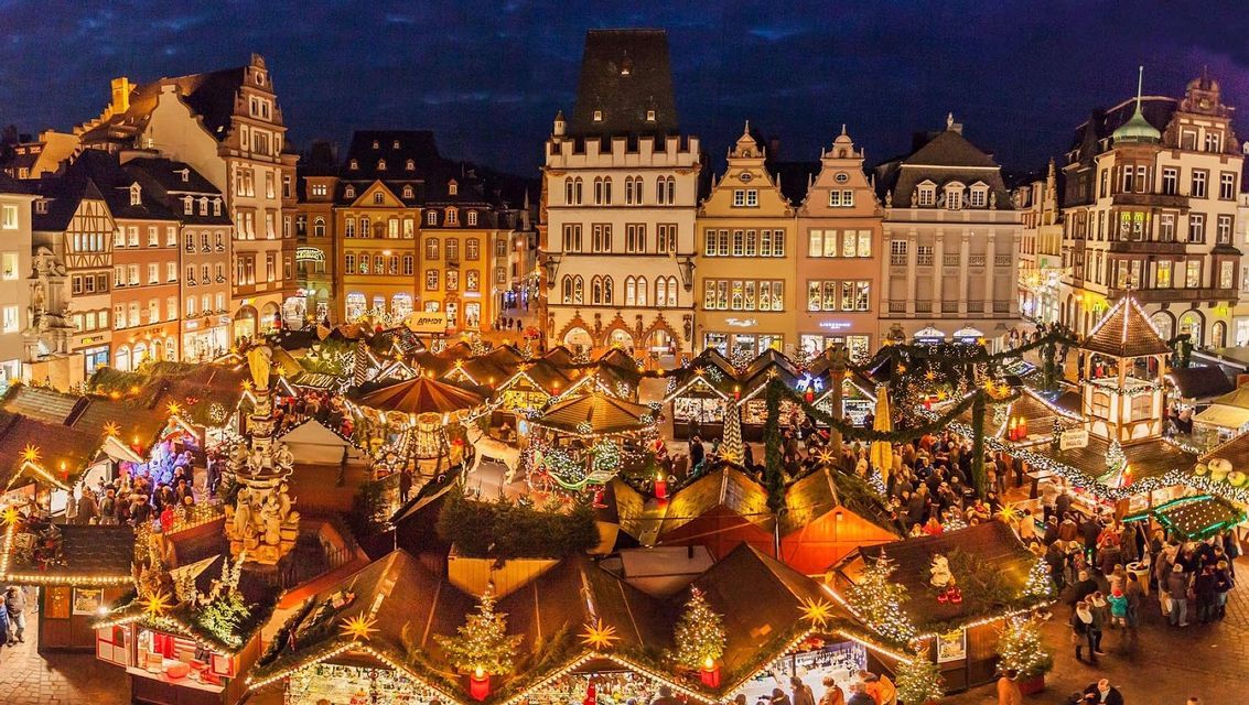 Una vista aerea di un affollato mercatino di Natale di notte, con bancarelle illuminate poste di fronte a storici edifici europei.