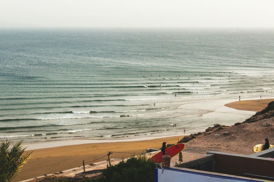 Vue plongeante sur une plage avec de nombreux surfeurs dans l'océan, et une personne tenant une planche de surf rouge au premier plan.