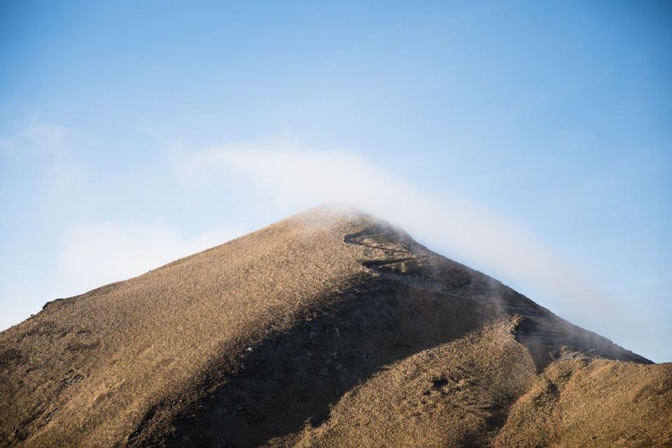 Un sentier sinueux serpente jusqu'à un sommet de montagne herbeux, tandis qu'un fin nuage flotte au-dessus du sommet sous un ciel bleu clair.
