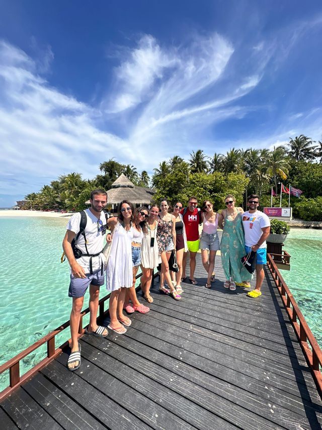 Un grupo de WeRoad posa para una foto en un muelle de madera sobre agua turquesa, con una playa tropical y palmeras al fondo.