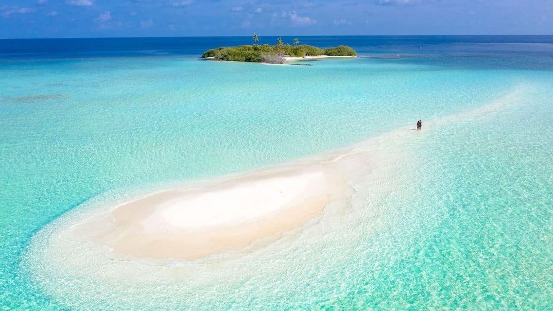 Una vista aérea de un banco de arena blanca rodeado de agua turquesa, donde dos personas caminan hacia una pequeña isla tropical.