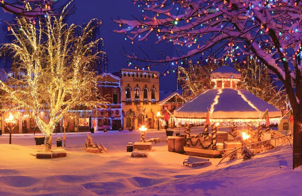 Una piazza cittadina innevata di notte, con edifici, alberi e un gazebo decorato con luci natalizie colorate.