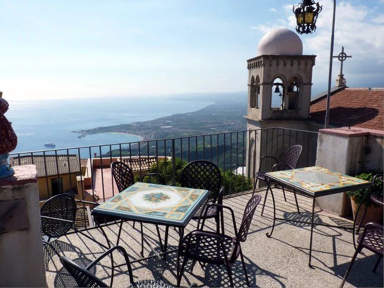 Tavoli e sedie a mosaico su una terrazza con vista su una cittadina costiera e sul mare, con un campanile in pietra in evidenza.