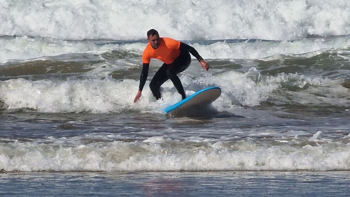 Ein Mann in einem orangefarbenen Hemd und schwarzem Neoprenanzug reitet eine Welle auf einem hellblauen Surfbrett.