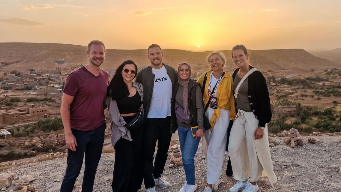 Eine WeRoad Gruppenreise von sechs lächelnden Personen auf einem Hügel mit Blick auf ein Dorf in einer trockenen Landschaft bei Sonnenuntergang.
