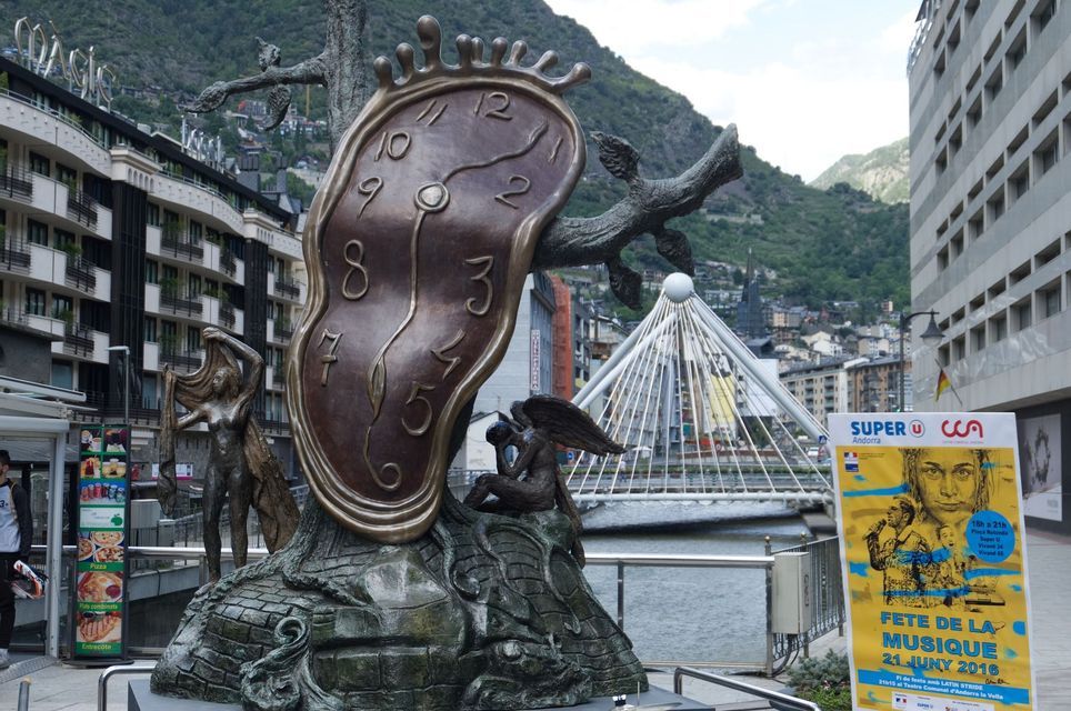 Una grande scultura in bronzo di un orologio che si scioglie su una strada cittadina, con un ponte bianco e montagne visibili sullo sfondo.
