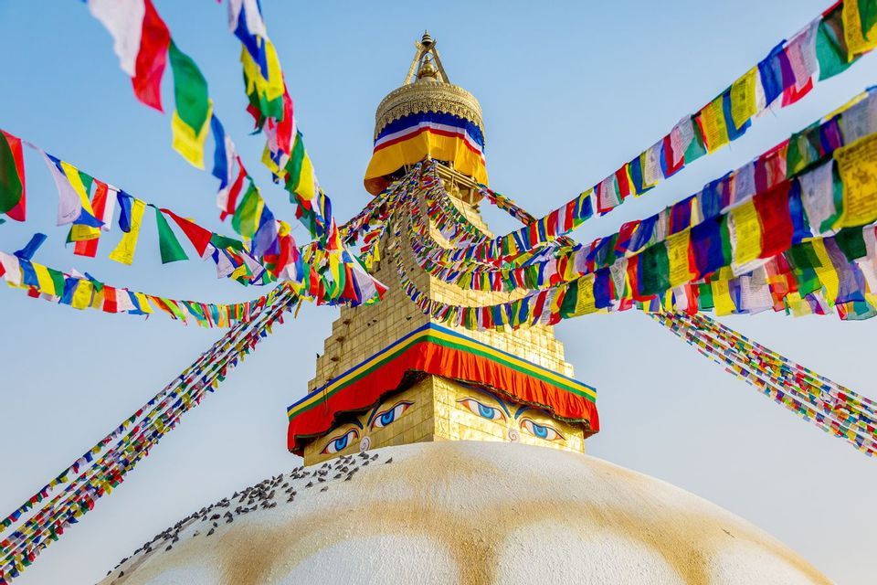 Bandiere di preghiera colorate sono appese alla guglia dorata di uno stupa buddista con occhi dipinti contro un cielo azzurro chiaro.