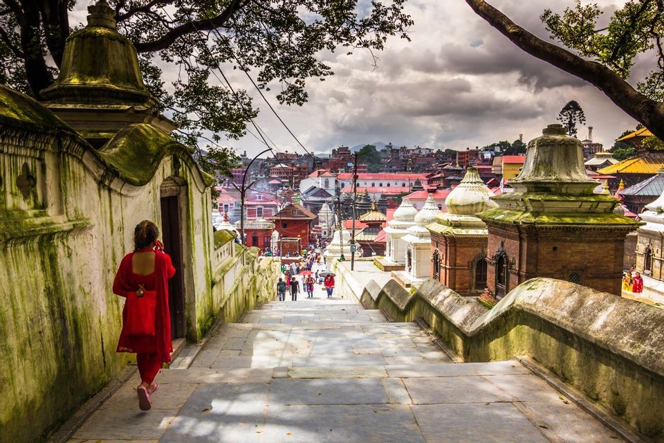Una donna in rosso scende ampie scale di pietra che si affacciano su una città di templi tradizionali sotto un cielo nuvoloso.