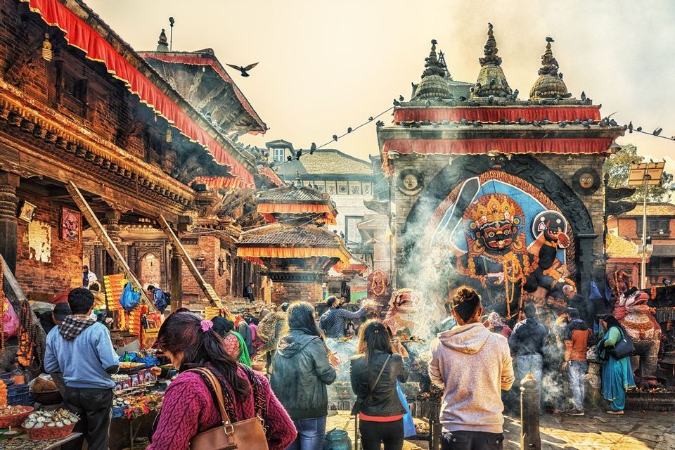 Una folla di persone si raduna in una vivace piazza del tempio con bancarelle del mercato, circondata da edifici ornati e un santuario di divinità fumante.