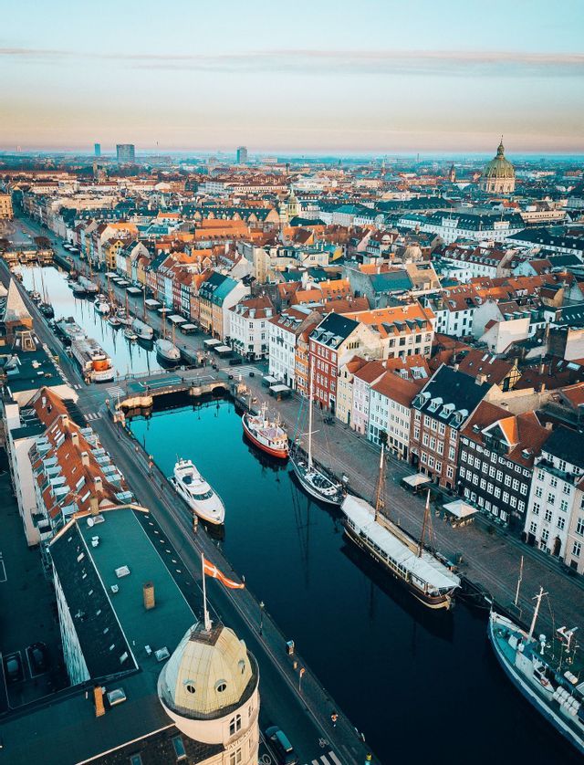 Vista aerea di un canale cittadino costeggiato da edifici colorati e barche, con una bandiera danese su una cupola in primo piano.