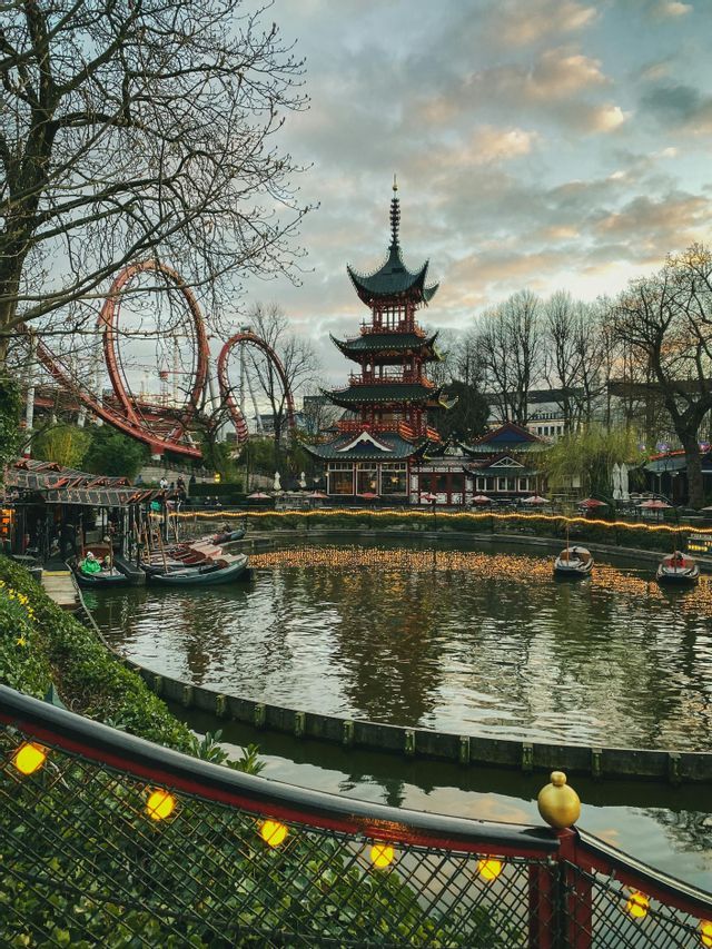 A Japanese pagoda and a red rollercoaster overlook a lake with boats at an amusement park during dusk.