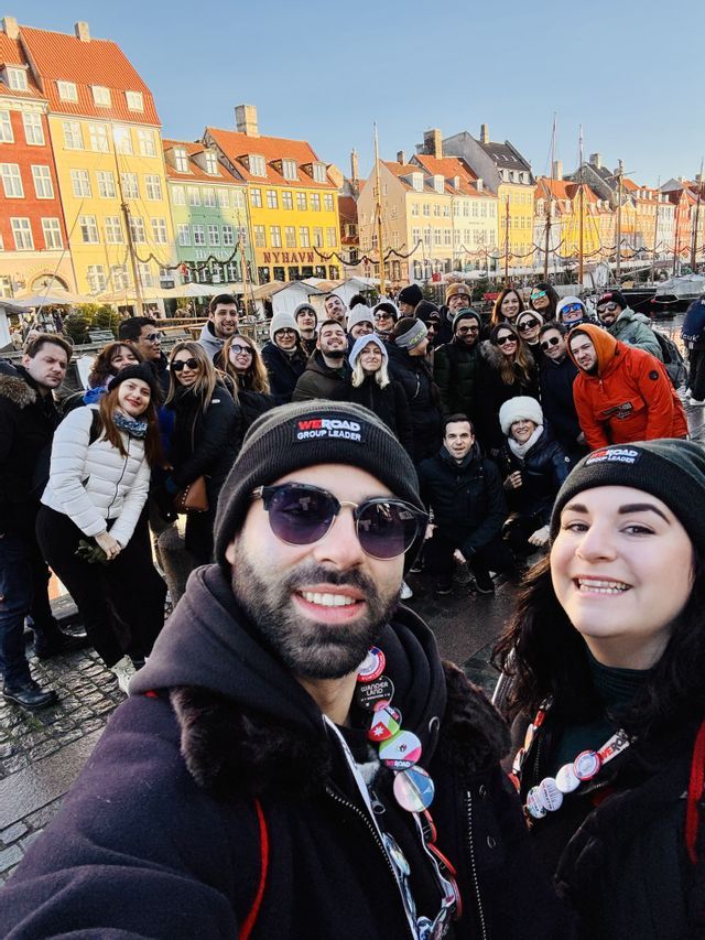 A WeRoad group trip takes a large group selfie on a cobblestone street in front of colorful canal-side buildings.