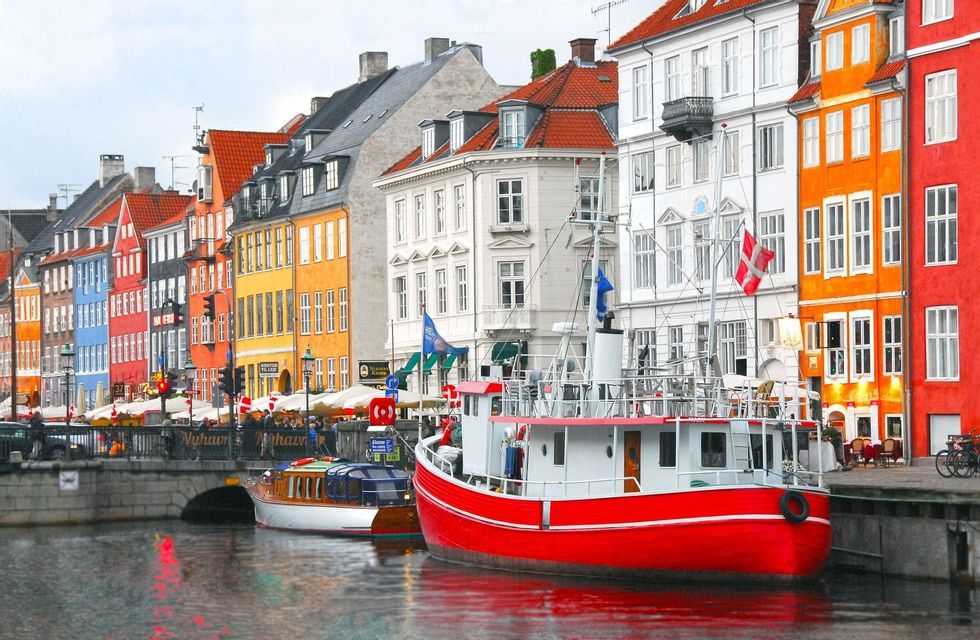A large red boat is docked in a canal, with colorful waterfront buildings and a small bridge in the background.