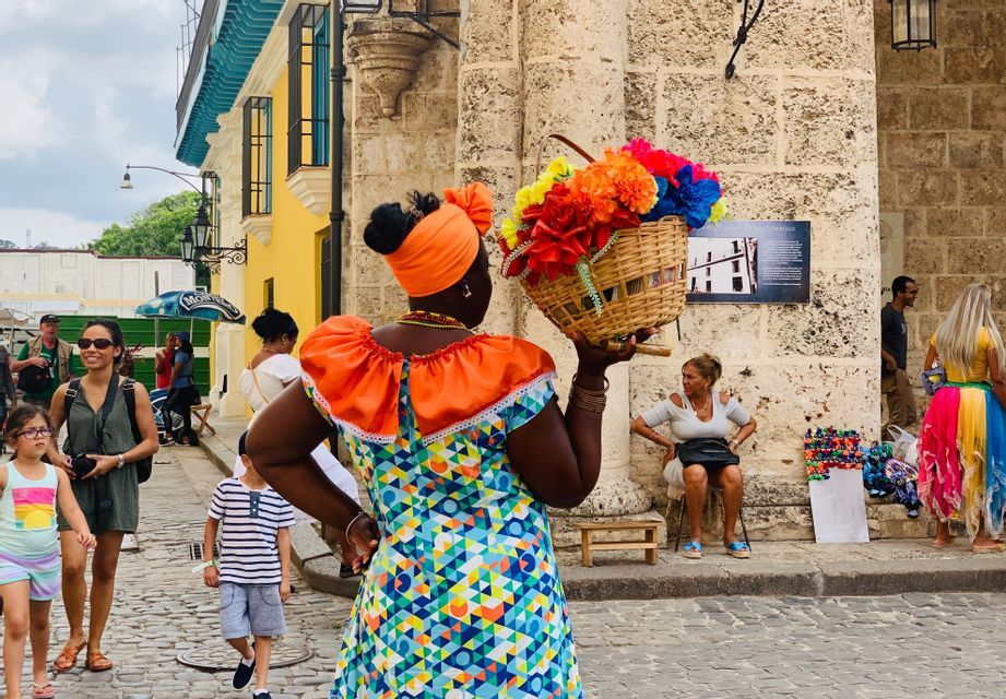 Una donna con un abito colorato a fantasia e un turbante arancione tiene un cesto di vivaci fiori di carta su una strada di ciottoli