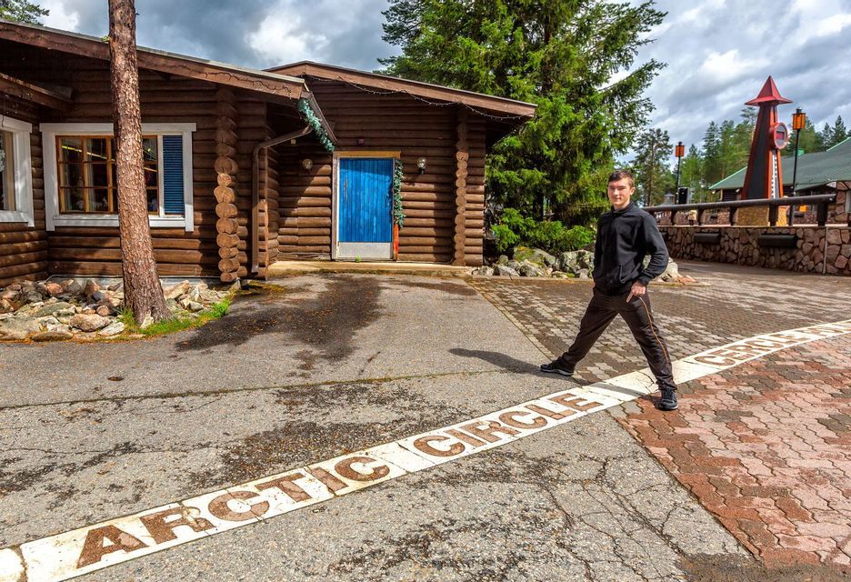 Ein junger Mann steht rittlings auf der am Boden gemalten Polarkreislinie vor einem Blockhaus.