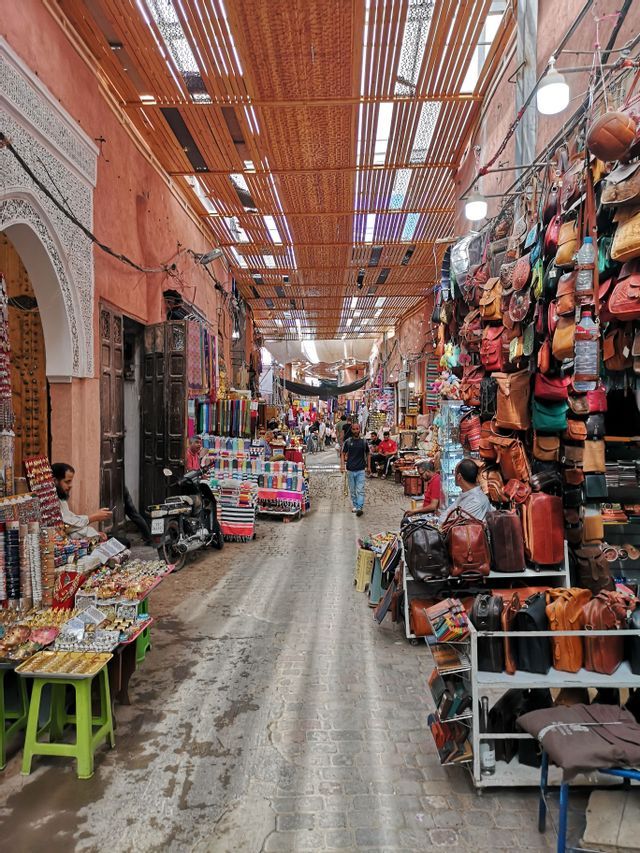 Eine belebte, enge Marktstraße mit Geschäften, die Ledertaschen und bunte Textilien unter einem Lamellendach anbieten.