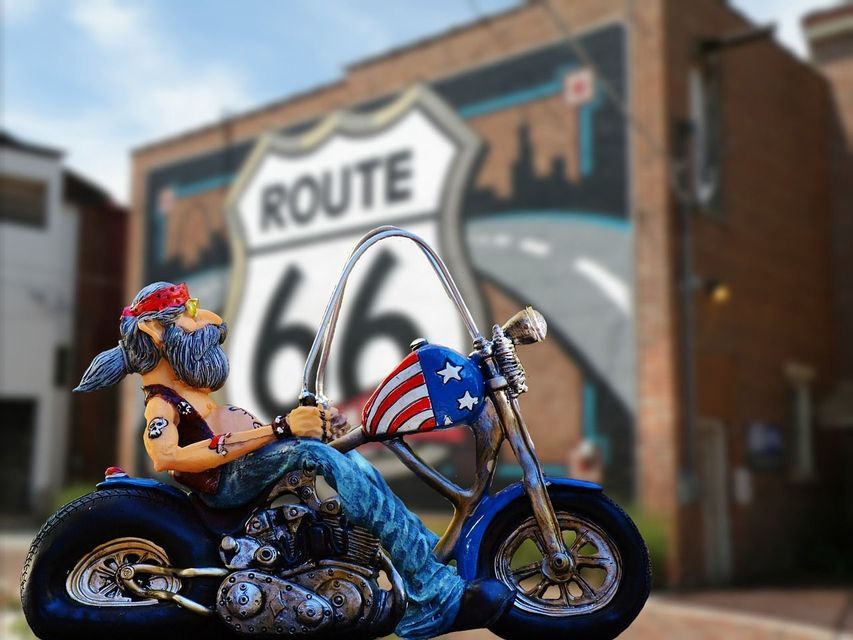 Una figura de un motociclista en una moto con el tanque de bandera estadounidense, frente a un mural difuminado del letrero de la Ruta 66 en una pared de ladrillo.