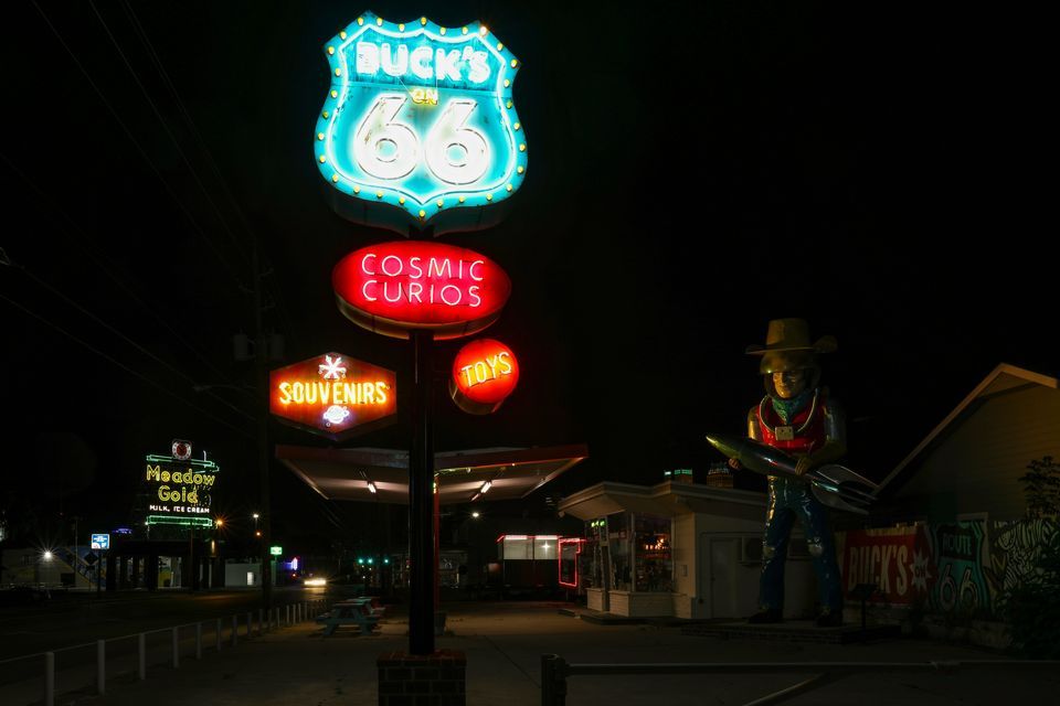 Coloridos letreros de neón de Buck's en la Ruta 66 y Cosmic Curios iluminan una atracción al borde de la carretera con una estatua de astronauta por la noche.