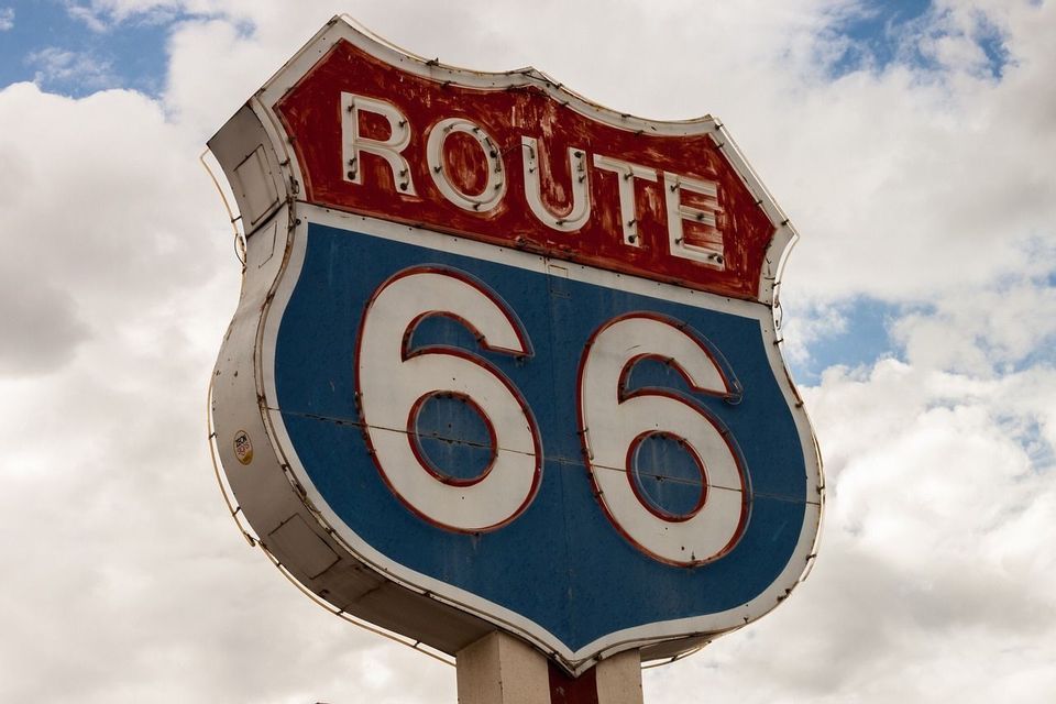 Una señal de carretera clásica y desgastada de la Ruta 66 con colores rojo, blanco y azul, sobre un cielo nublado.