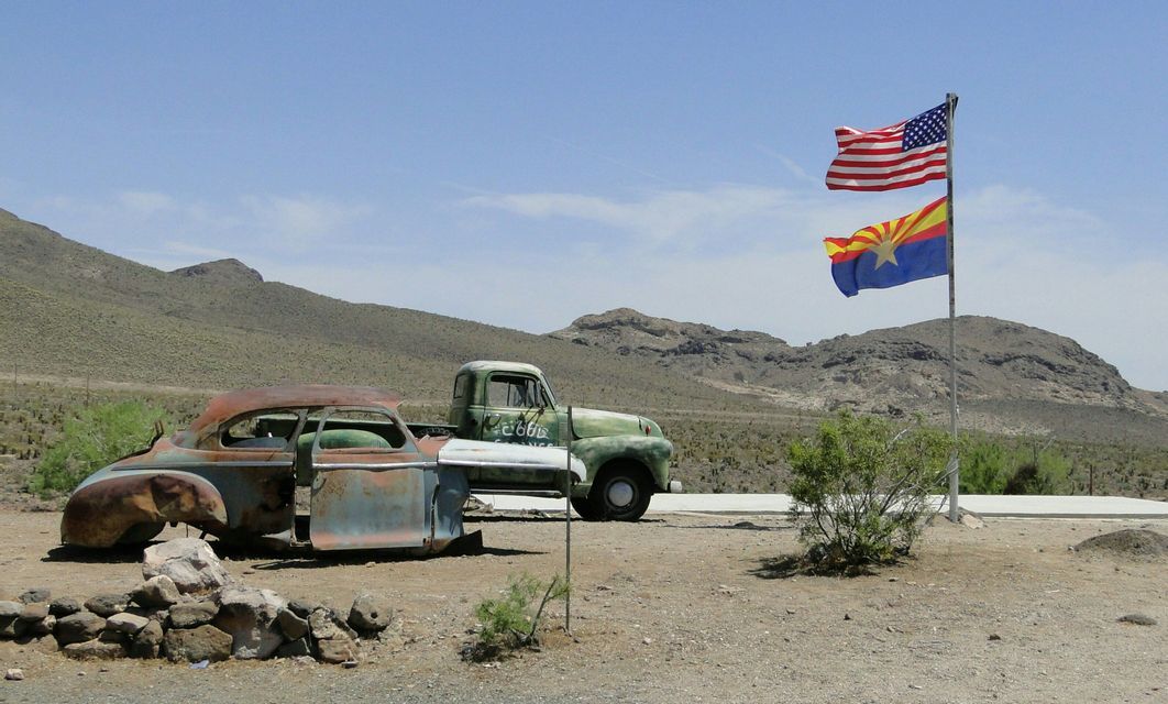 Un coche oxidado y una camioneta antigua estacionados en un paisaje desértico con las banderas de Estados Unidos y Arizona ondeando en un mástil.