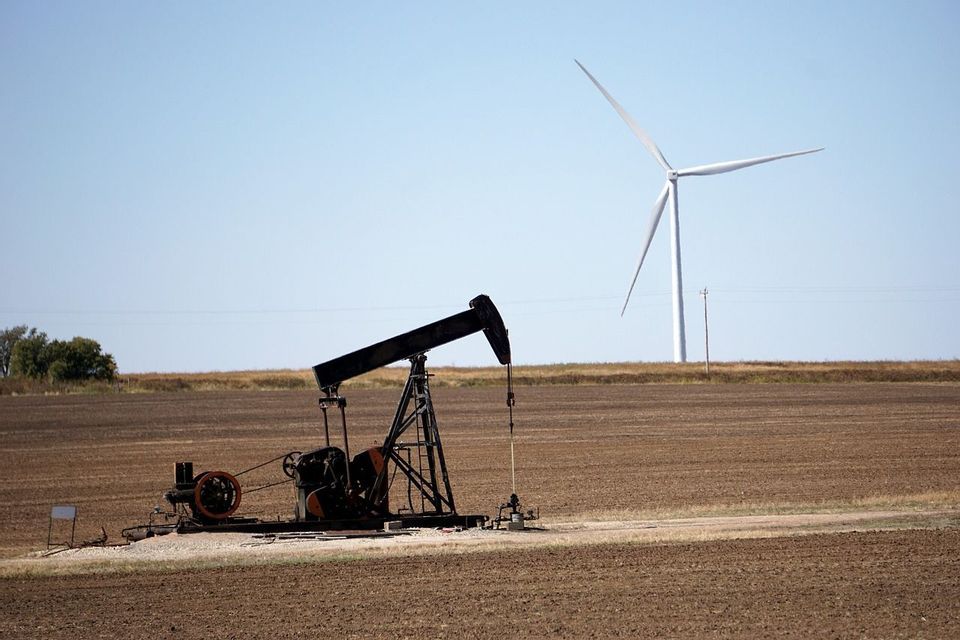 Un balancín petrolero en un campo arado con una turbina eólica al fondo bajo un cielo azul despejado.