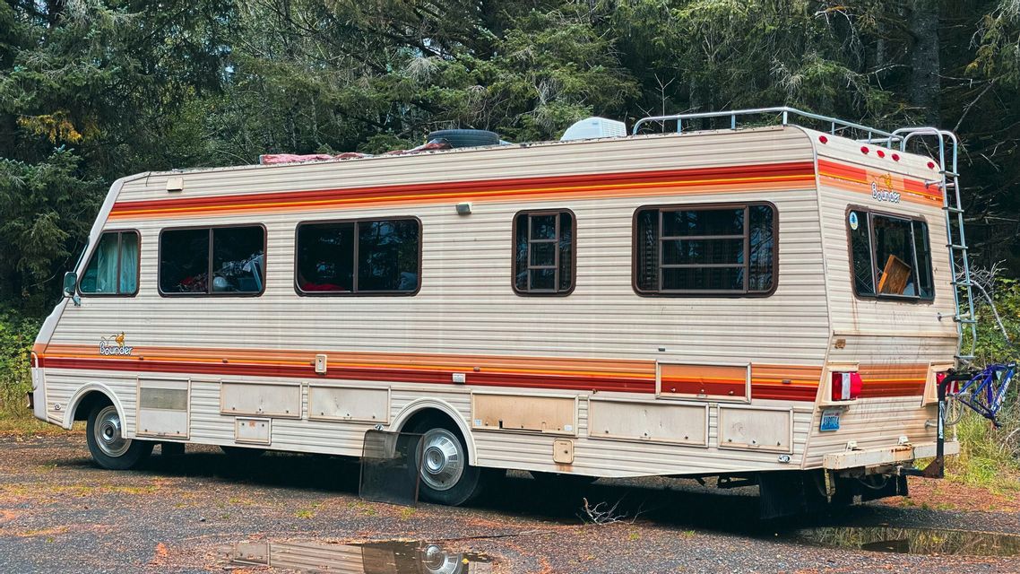 Una autocaravana grande, modelo antiguo, con rayas naranjas y rojas está estacionada en un terreno de grava junto a un bosque.
