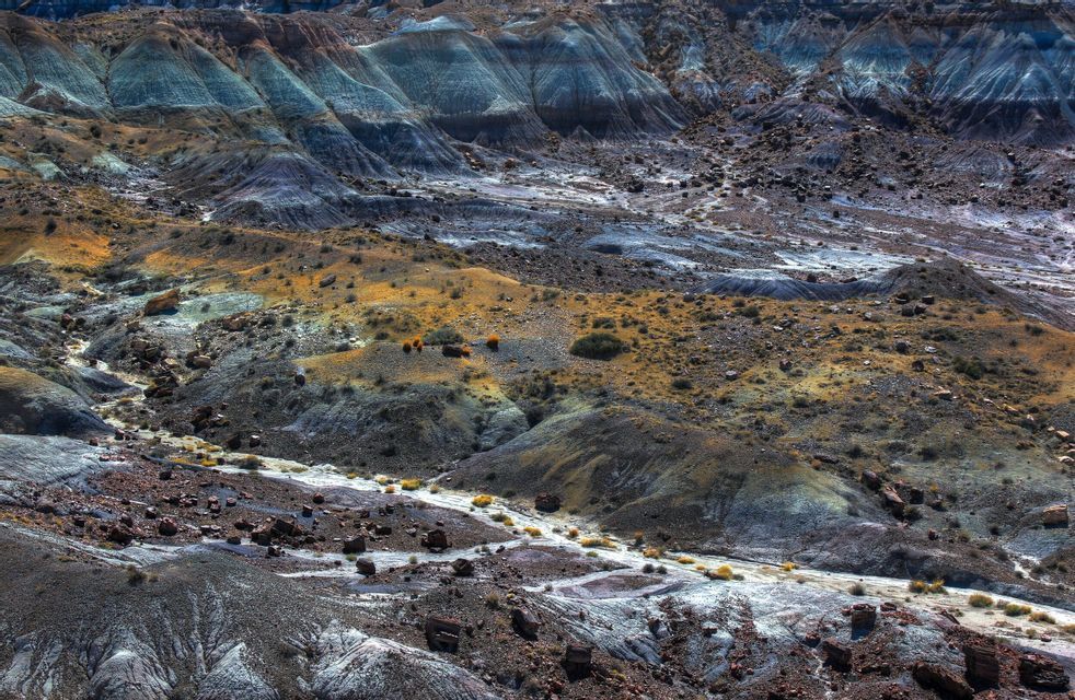 Un vasto paisaje de tierras baldías erosionadas con estratos coloridos de sedimento azul, gris y amarillo bajo la luz brillante del día.