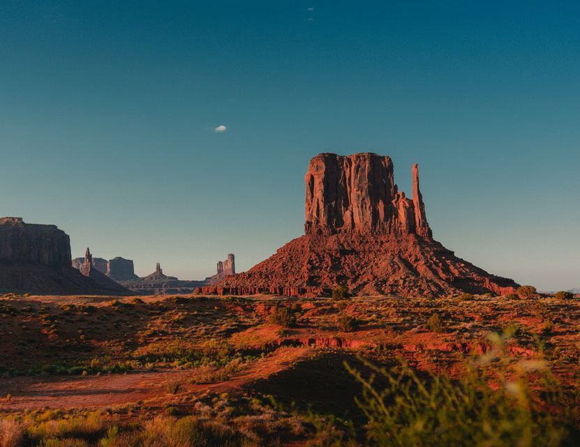 Una gran mesa roja y otras formaciones rocosas se alzan en un paisaje desértico a la hora dorada bajo un cielo azul claro.