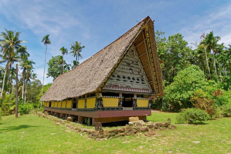 Una longhouse tradizionale con tetto di paglia e facciata dipinta si erge in una radura erbosa circondata da palme.