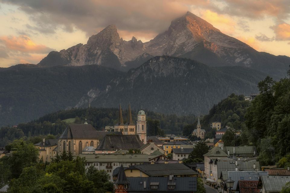 Ein Alpendorf mit markanten Kirchtürmen liegt bei Sonnenuntergang am Fuße hoch aufragender, felsiger Berge.