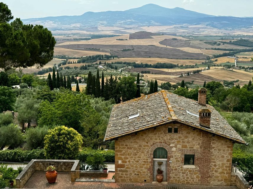 Una casa in pietra con tetto in tegole si affaccia su una vasta valle rurale con campi a patchwork ondulati e montagne sullo sfondo.