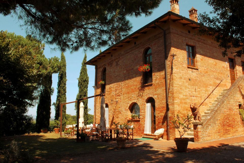 Una casa rustica in mattoni rossi con patio esterno e cipressi, bagnata dalla luce del sole pomeridiano.