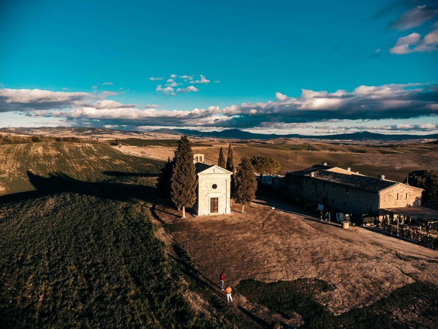 Veduta aerea di una cappella in pietra e un casale su una collina, circondati da campi ondulati verdi e marroni sotto un cielo blu.
