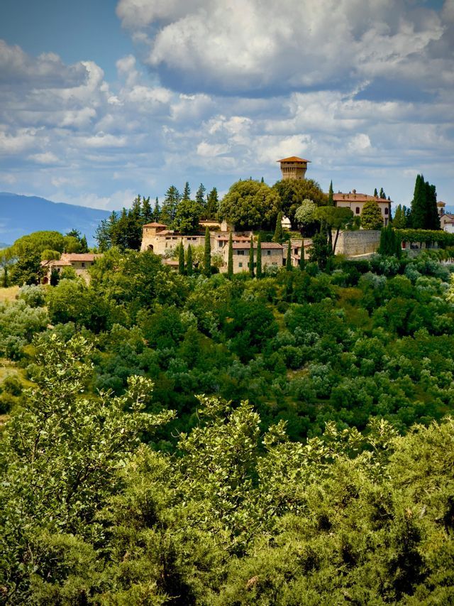 Un borgo in pietra con una torre prominente è immerso tra gli alberi su una rigogliosa collina verde sotto un cielo parzialmente nuvoloso.