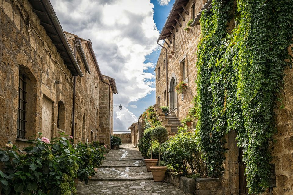 Uno stretto vicolo di ciottoli si inerpica tra vecchi edifici in pietra, uno dei quali è coperto di edera verde, sotto un cielo parzialmente nuvoloso.
