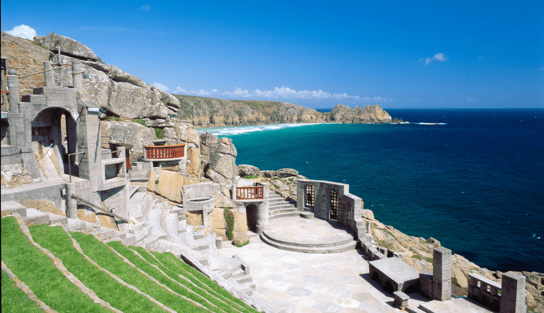 Un anfiteatro in pietra con gradinate in erba costruito in una scogliera rocciosa, affacciato su un oceano blu calmo sotto un cielo sereno.