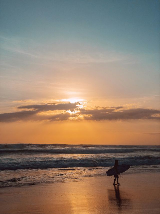 Un surfista en silueta camina por una playa mojada llevando una tabla de surf al atardecer.