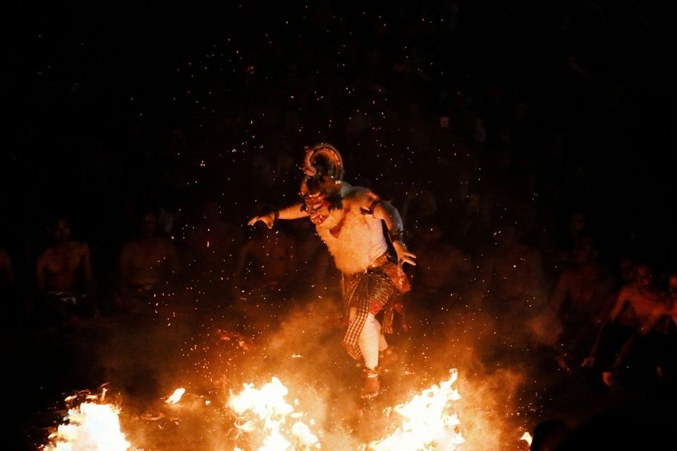 Un artista enmascarado con vestimenta tradicional salta sobre una hoguera, esparciendo chispas en la oscuridad.