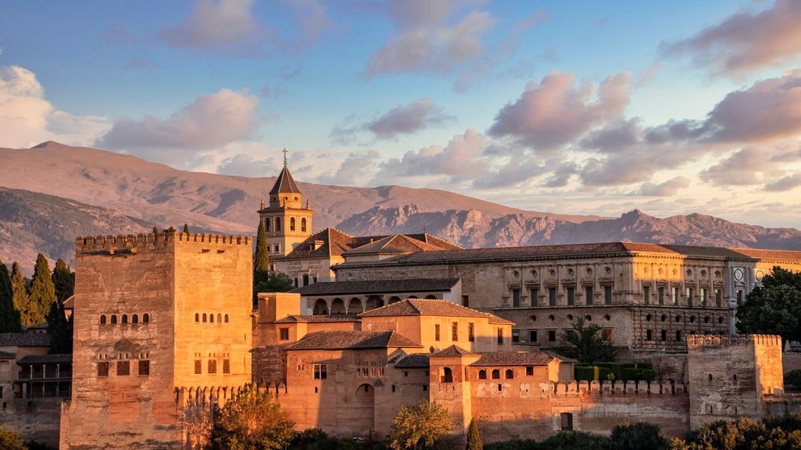 Un complejo de fortaleza de piedra histórico está bañado en luz dorada al atardecer, con una cordillera al fondo.